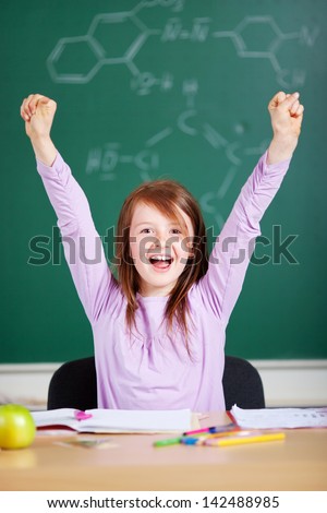 Jubilant excited little girl in school cheering and raising her arms in the air against a blackboard background