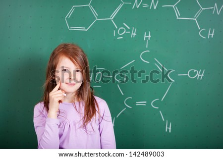 Pretty female student puzzling a chemistry question standing against a blackboard with a drawing of a chemical formula with a thoughtful expression