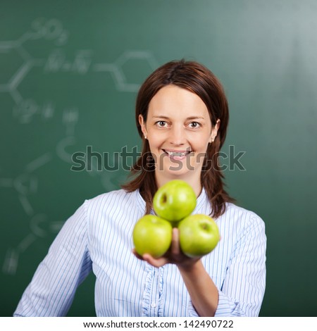 Smiling female teacher standing in front of the blackboard with a handful of fresh green apples