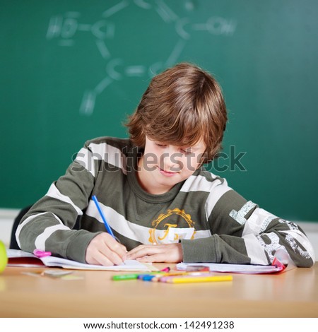 Young schoolboy sitting at his desk in front of the blackboard writing notes