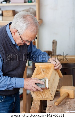 Senior male carpenter using sandpaper for polishing birdhouse at worktable in workshop