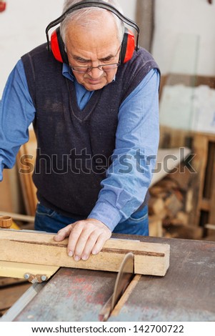 Senior man wearing red ear protectors while using table saw for cutting wood at workshop