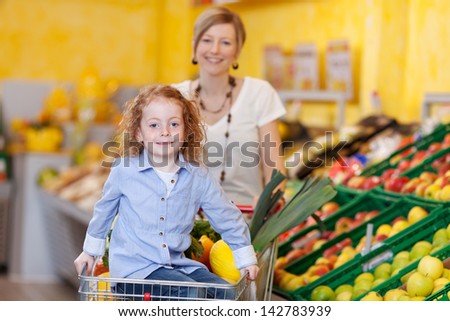 Pretty little girl and her mother in a supermarket pushing a trolley filled with fresh fruit and veg with the daughter riding on the front