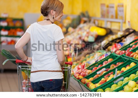 Blond woman shopping for groceries in a supermarket standing with her back to the camera pushing her trolley past the fresh produce