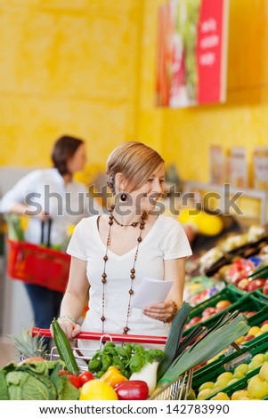 Happy young woman shopping vegetables and fruits while holding checklist in grocery store