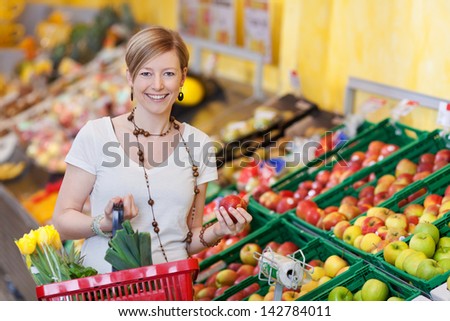 Happy woman buying fresh produce in a supermarket standing with a basket of groceries in one hand and a ripe red apple in the other