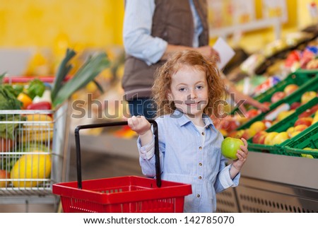 Portrait of little girl holding shopping basket and apple with woman in background at grocery store