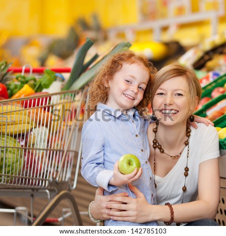 Portrait of happy little girl holding apple while mother embracing her in grocery store