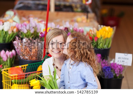 Cute little girl with curly red head kissing her mother as they shop for fresh flowers in a supermarket