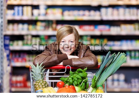 Woman With A Shopping Cart Of Fresh Produce Resting Her Chin On Her Hands As She Leans On The Cart Smiling At The Camera