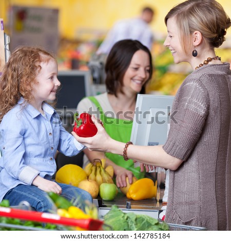 Happy Young Woman Giving Capsicum To Daughter With Cashier Smiling In Background At Super Market