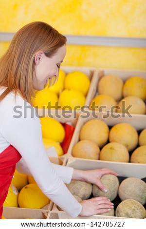 Attractive young female sales assistant repacking melons in a display in a fresh food supermarket with assorted melons displayed in boxes