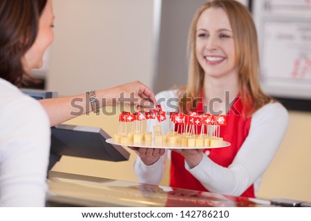 Happy saleswoman offering small cheese blocks with Swiss flag to customer in grocery store