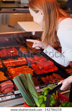Woman purchasing meat at a delicatessen leaning forwards pointing to her selection behind a glass counter display