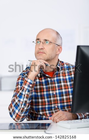 Portrait of a mature balding caucasian man wearing glasses, sitting at the office desk, holding his chin with the hand and looking up and away in a serious and pensive mood