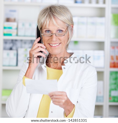 Portrait of happy senior pharmacist using cordless phone while holding prescription paper in pharmacy