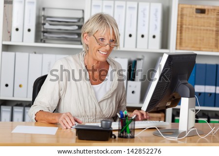 Tech savvy elderly woman working on the computer and smiling, sitting inside her office.
