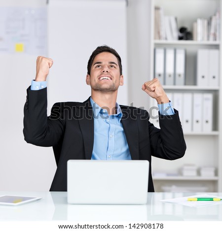 Successful businessman rejoicing raising his face to the sky and punching the air with his fists as he sits at his desk in the office
