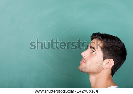 Head profile portrait of a thoughtful young male student in front of a green blackboard looking up towards copyspace on the board
