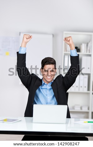 Jubilant businessman sitting in front of his open laptop with his fists raised in the air and a beaming smile of success