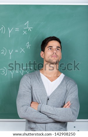 Thoughtful man standing with his arms folded in front of the chalkboard in a classroom with mathematical equations staring off into the distance