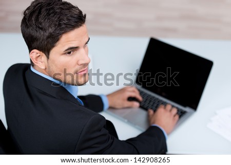 High angle over the shoulder view of a determined handsome young businessman typing on his laptop computer with the blank screen visible to the camera