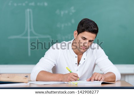 Handsome young man in university sitting at his desk in front of the blackboard working on his notes with a luminous highlighter