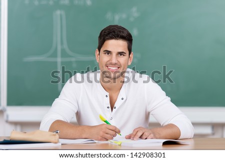 Happy handsome young male university student sitting in front of a blackboard at his desk taking notes looking up at the viewer with a lovely smile