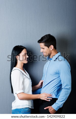 Beautiful young couple in love standing next to each other and looking into each others eyes, isolated on blue background.