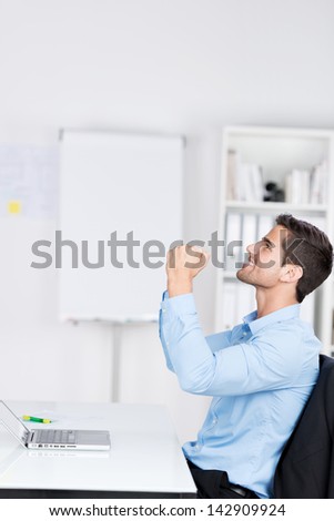 Side view of happy young businessman with clenched fists celebrating victory at desk in office