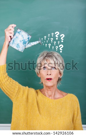 Senior female teacher holding watering can with question marks on chalkboard representing confusion