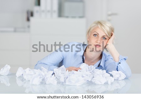 Young thoughtful female executive sitting with crumpled paper balls at desk looking in camera