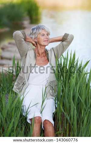 Happy senior woman with hands behind head relaxing by lakeside in park