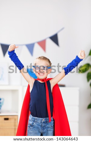 Portrait Of Happy Little Boy With Arms Raised In Super Hero Costume At Home