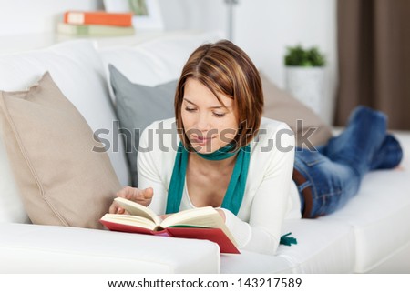 Woman reading a book on the couch lying on her stomach facing towards the camera