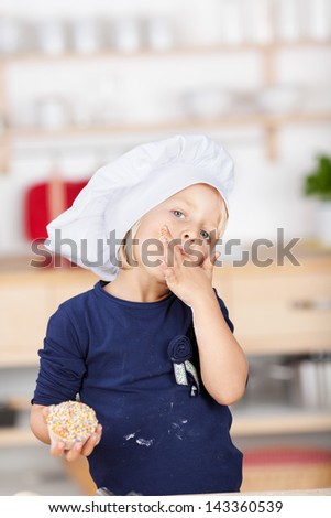 Cute small chef in an over sized chefs toque standing looking at the camera licking her fingers while cooking in the kitchen