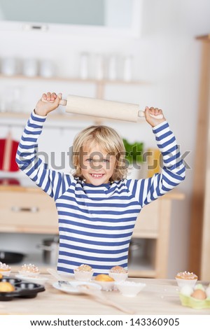 Smiling little girl with a wooden rolling pin grinning and holding it above her head as she stands in the kitchen baking cupcakes