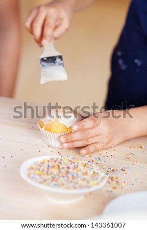 Child decorating a cupcake glazing the top with icing using a brush before adding sprinkles