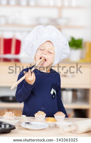 Playful little girl baking cupcakes standing at a kitchen counter in her chefs hat happily licking a wooden spoon, while tasting the mixture