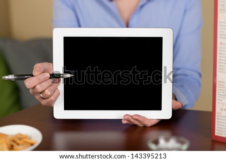Woman pointing to a blank tablet display with a pen on a coffee shop with a cup of snacks and menu on the side
