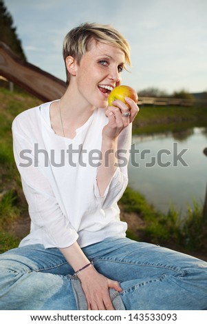 Portrait of a smiling young beautiful woman eating an apple by the country lake