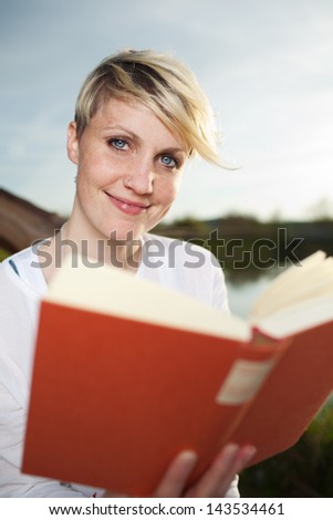 Smiling young beautiful young blond woman reading a book by the country lake