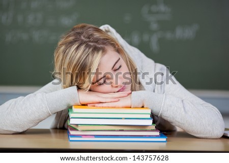 Bored female student sleeping on stacked books at desk in classroom