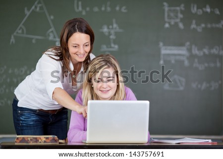 Female teacher assisting student in using laptop at desk in classroom
