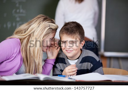 Schoolgirl whispering to her male friend in the classroom while the teacher is writing on the blackboard.