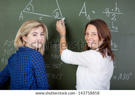Portrait of happy teacher writing on chalkboard while standing with student in classroom