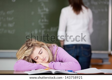 Bored female student sleeping at desk with teacher in background at classroom