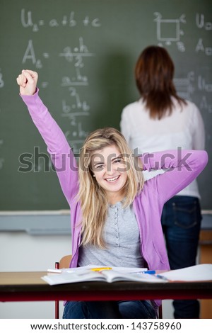 Portrait of happy female student stretching at desk with teacher in background at classroom