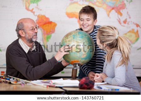 Senior male teacher pointing at globe while students looking at it at desk in classroom
