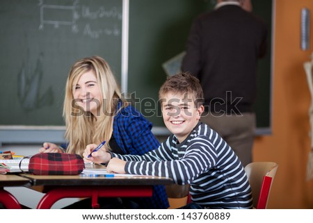 Portrait of happy classmates sitting at desk with teacher writing on blackboard in classroom